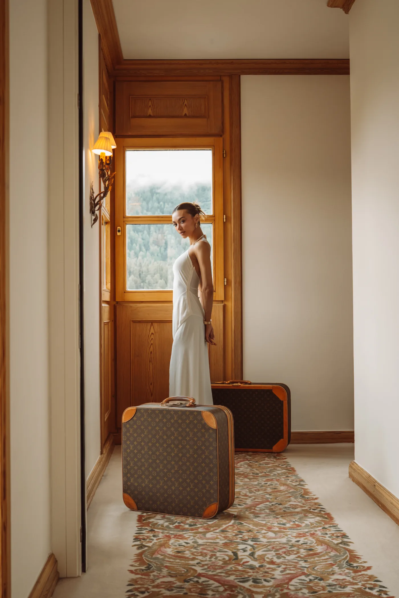 Kylie Verzosa in a white gown in a wood-panelled corridor with monogrammed luggage